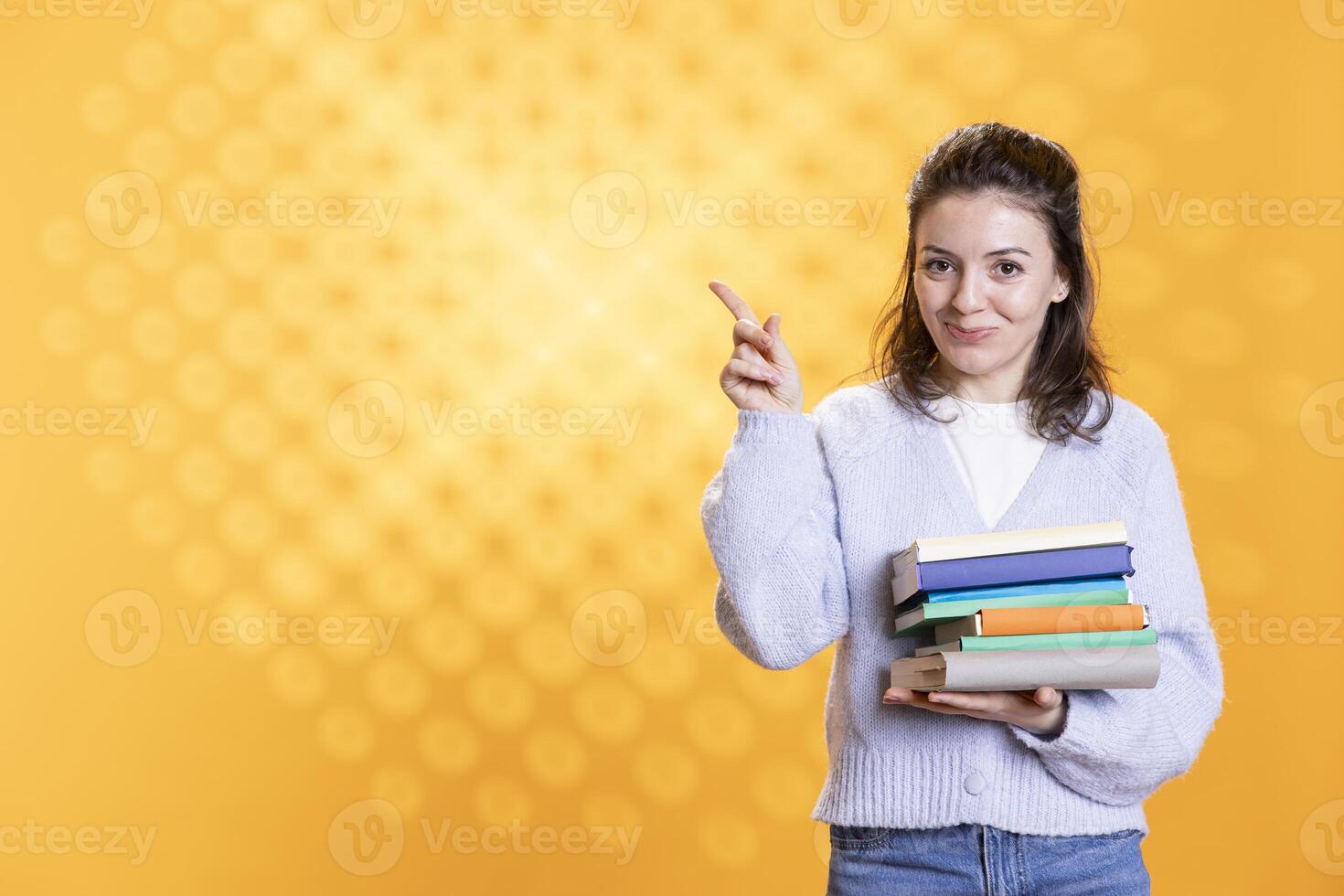 Portrait of radiant woman with stack of books in hands pointing towards empty copy space, isolated over studio background. Happy bookworm holding pile of novels, pointing finger to message photo
