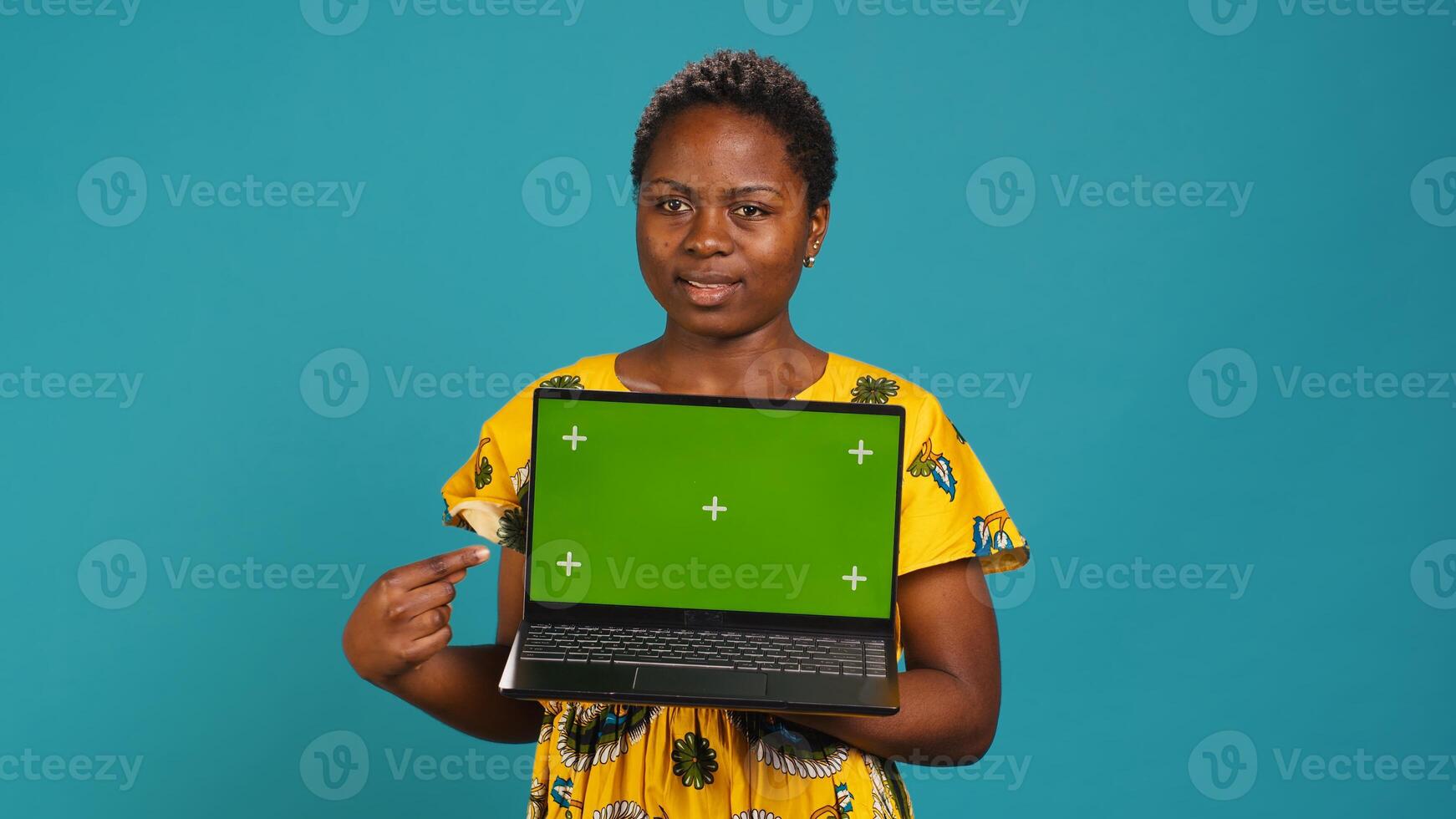 Young adult showing a laptop with isolated mockup screen in studio, presenting a display with green screen against blue background. Female model posing with chroma key. Camera A. photo