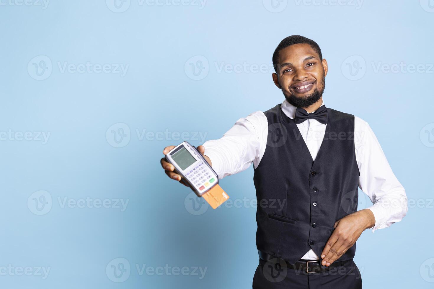 Professional hotel porter presenting the pos terminal and credit card, symbolizing the payment at front desk against blue background. Receptionist with tie asking for the transaction. photo