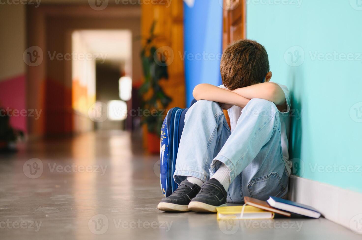 Little boy sitting alone on floor after suffering an act of bullying. Sad young schoolboy sitting on corridor with hands on knees and head between his legs photo