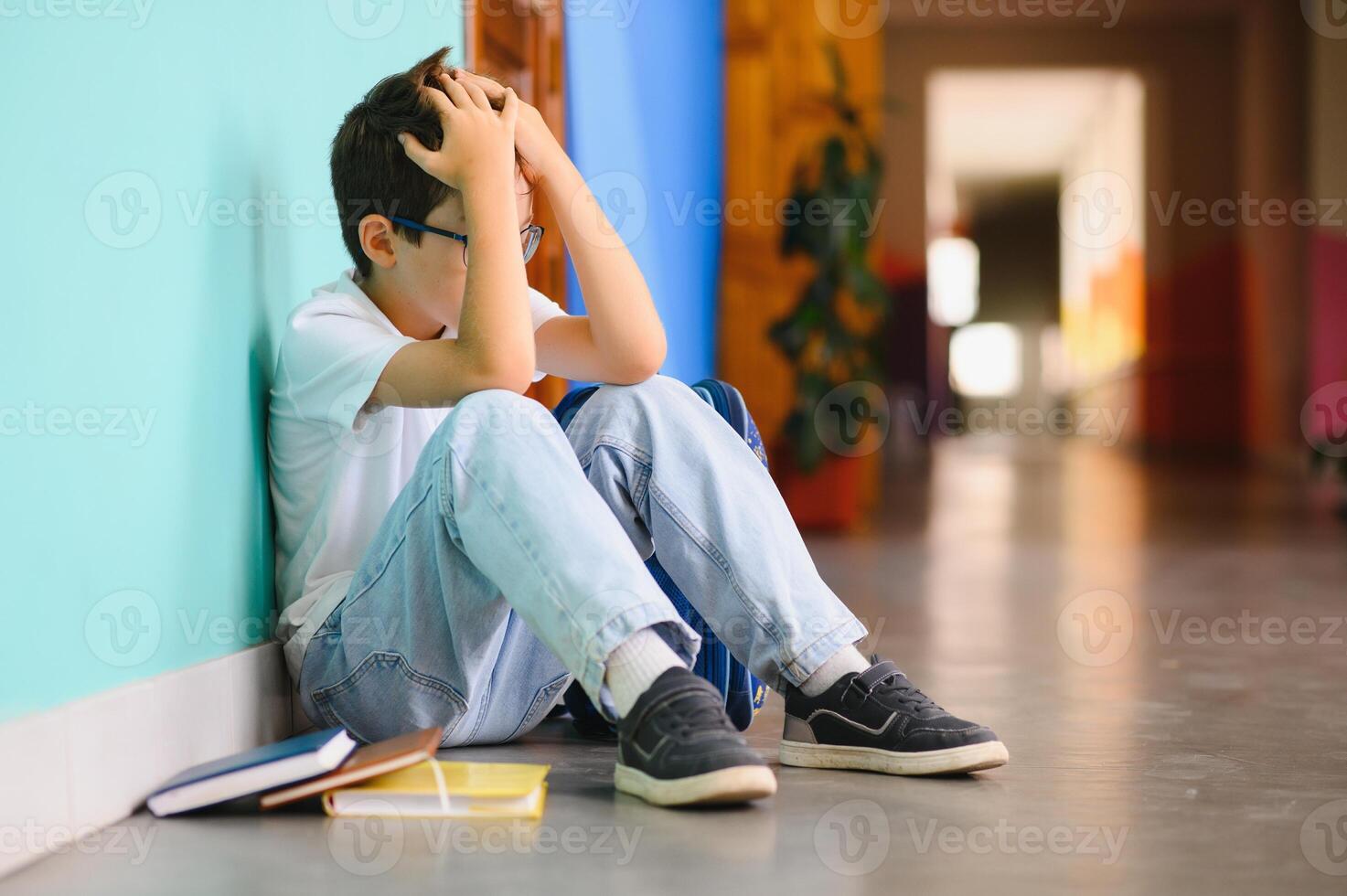 Little boy sitting alone on floor after suffering an act of bullying. Sad young schoolboy sitting on corridor with hands on knees and head between his legs photo