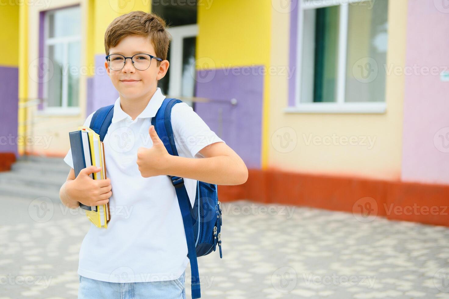 Portrait of little schoolboy in glasses and with backpacks standing on the school building. Study concept photo