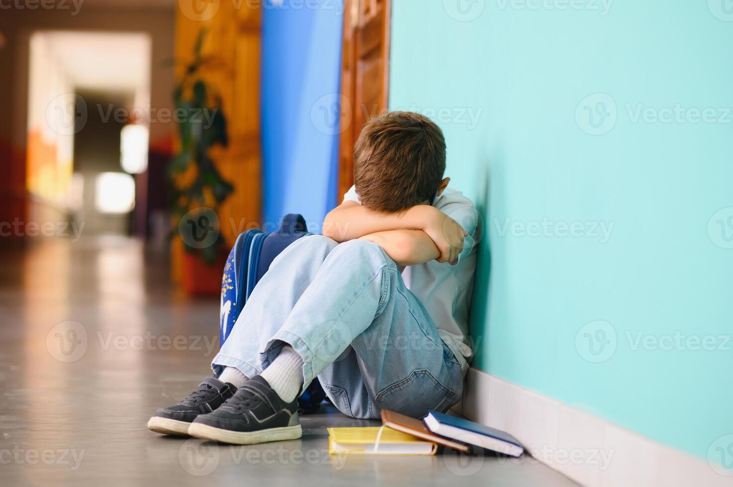 Little boy sitting alone on floor after suffering an act of bullying. Sad young schoolboy sitting on corridor with hands on knees and head between his legs photo
