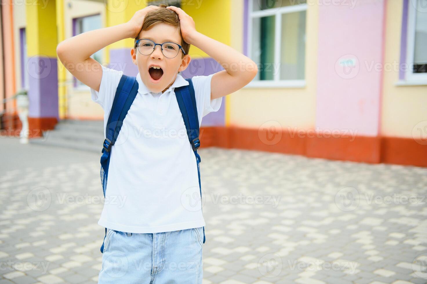 Portrait of little schoolboy in glasses and with backpacks standing on the school building. Study concept photo