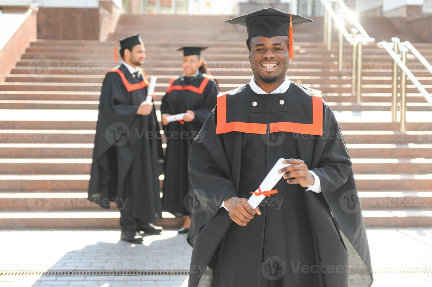 Happy graduate. Happy African man in graduation gowns holding diploma and smiling while his friends standing in the background photo