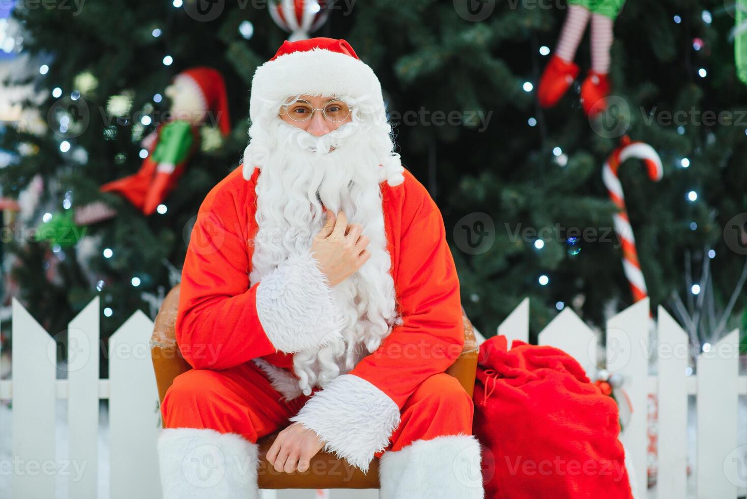 Santa Claus sitting on the background of the Christmas tree in the lobby of the modern shopping mall. Bokeh basic background for design. photo