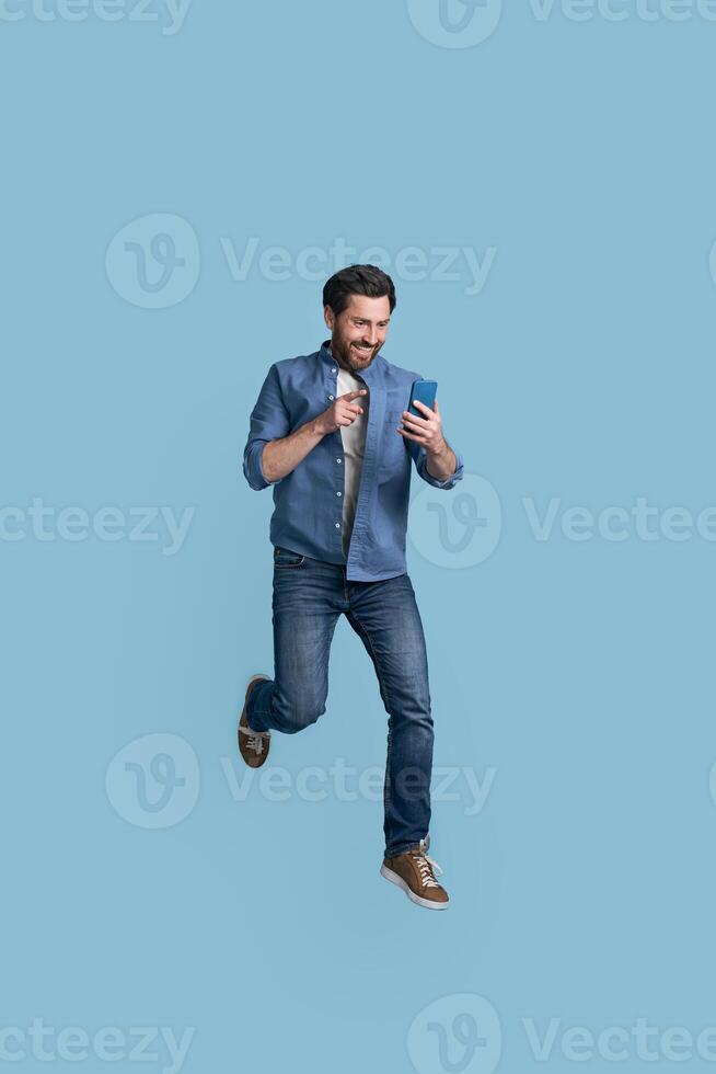 Excited man jumping and pointing at smartphone screen on blue background photo
