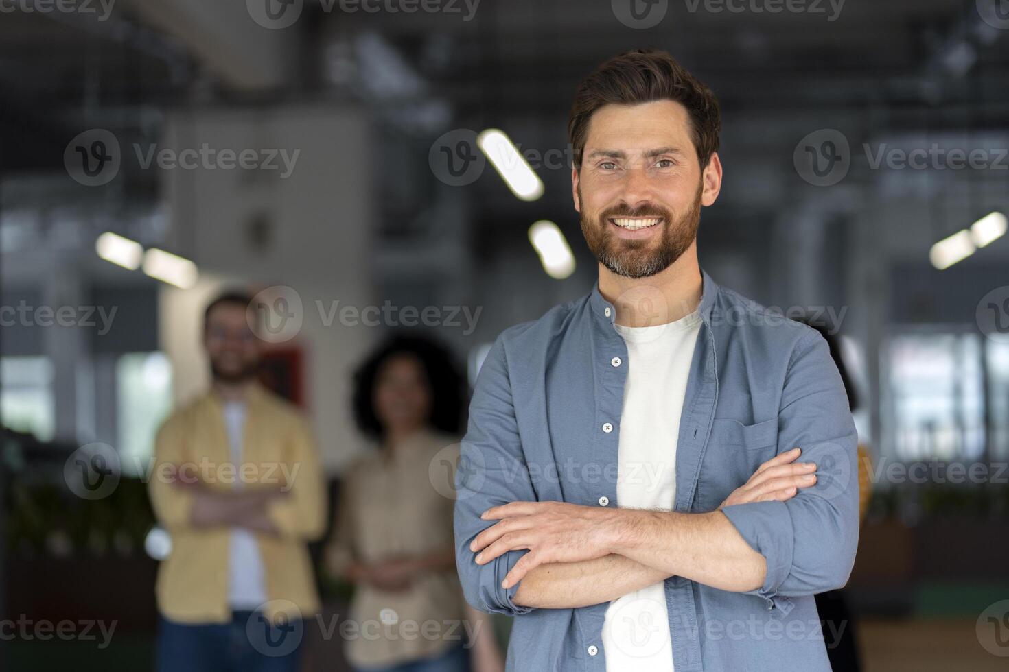 Successful businessman smiling with folded arms in office with coworkers in background photo