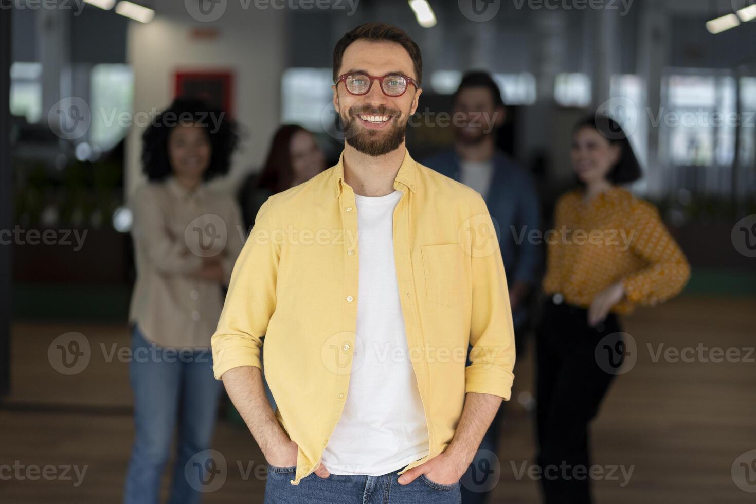 Confident businessman smiling with hands in pockets in office with team in background photo