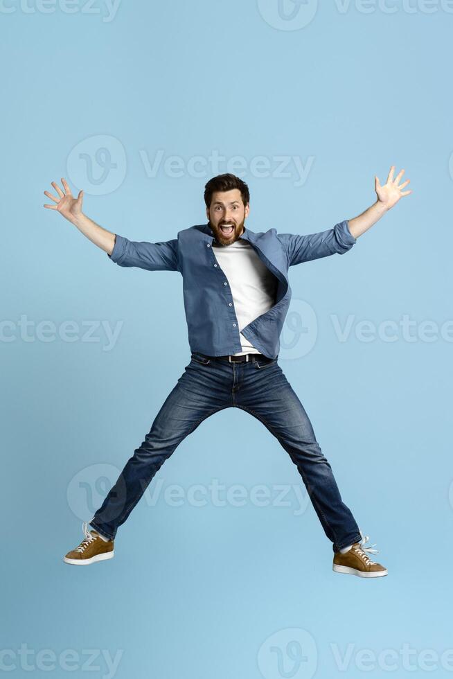 Excited man jumping with open mouth and spread arms on blue background photo