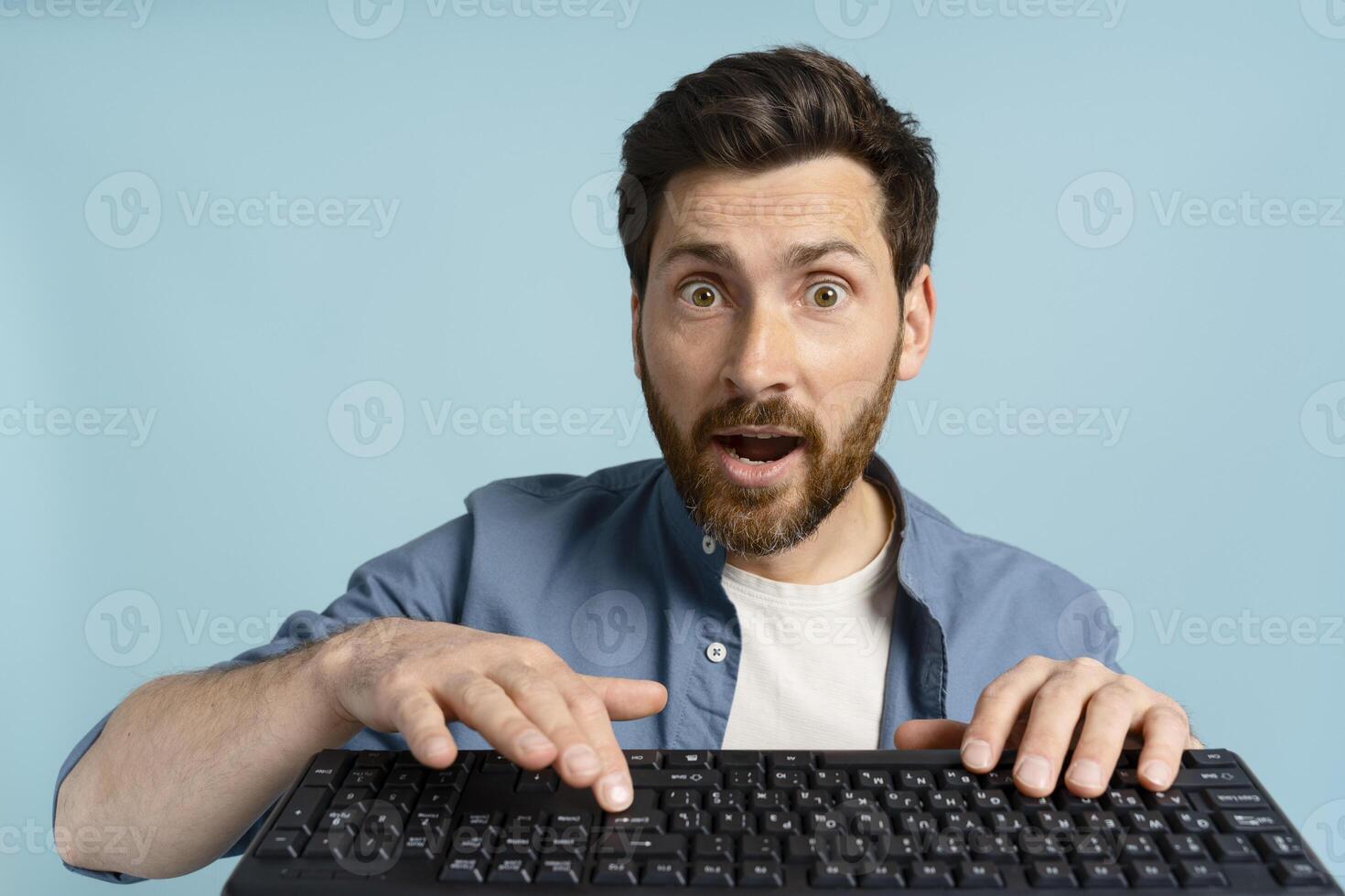 Shocked man looking at camera while typing on keyboard on blue background photo