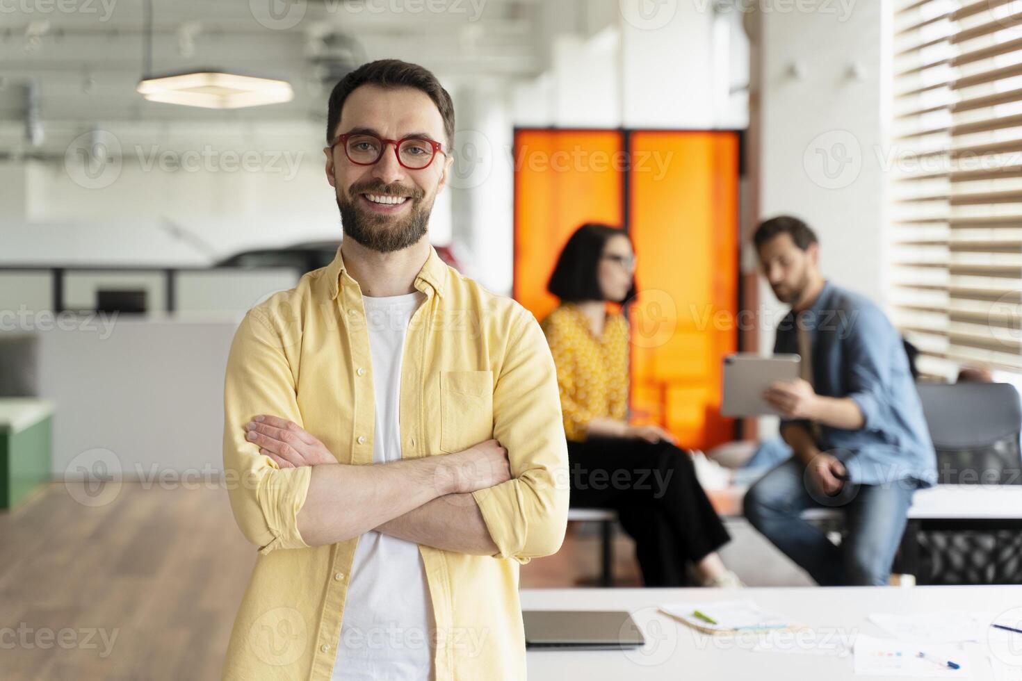 Confident businessman smiling with folded arms in busy office photo
