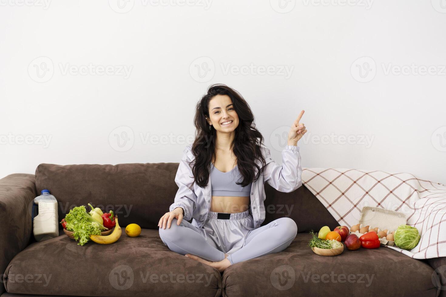 Young woman pointing up and showing healthy food on couch at home photo