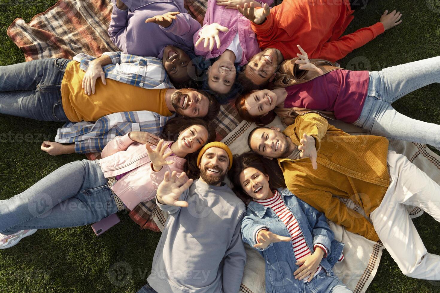 Diverse group of young people lying in circle on blanket and waving at camera photo