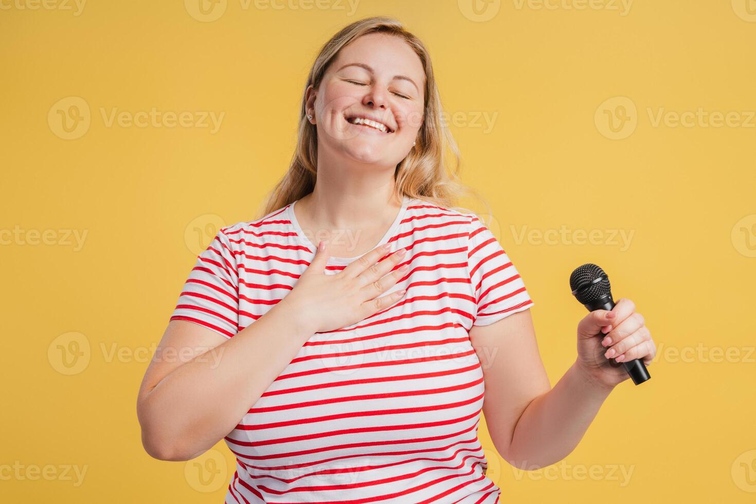 Smiling plus size woman enjoying singing holding hand on chest in studio photo