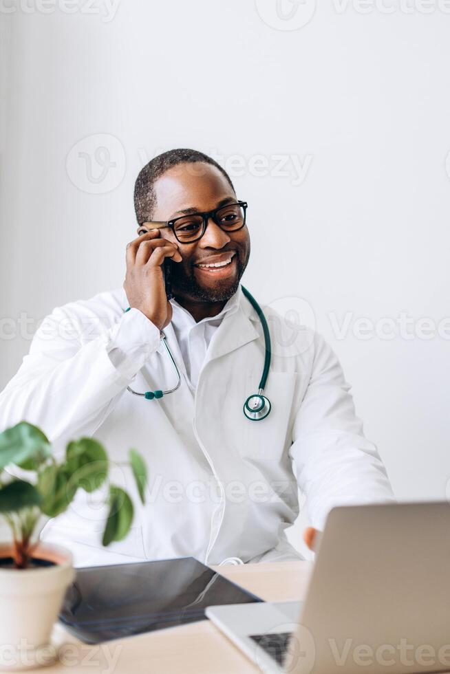 Smiling African American doctor in eyeglasses working with laptop talking on phone in cabinet photo
