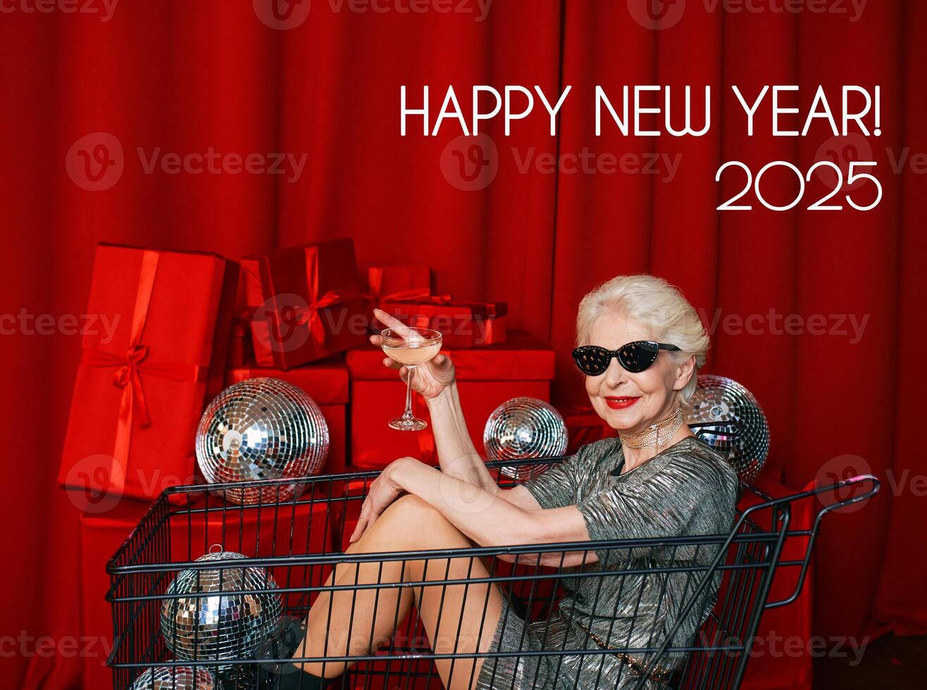 A stylish senior woman with gray hair, wearing sunglasses and a silver dress, sits in a supermarket cart at a party, sipping wine against a backdrop of red curtains. Happy New Year 2025 photo