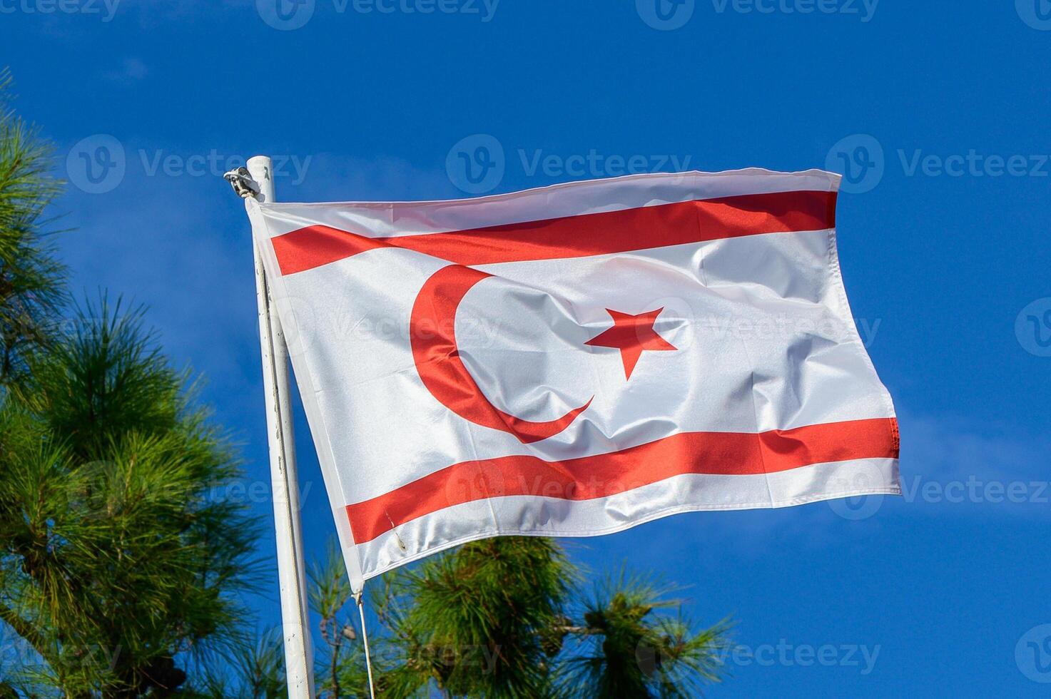The distinctive flag of North Cyprus billows against a bright blue sky near coastal pine trees, capturing a moment of serene national pride photo