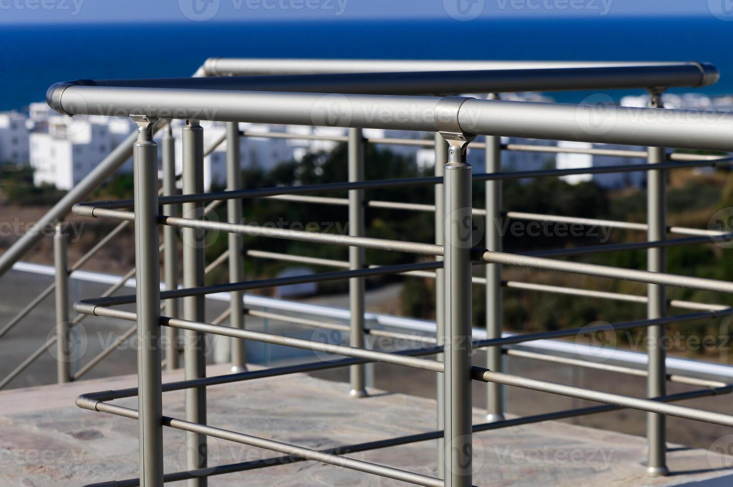 A Sleek Metal Railing Overlooking a Serene Coastal View With White Buildings Dotted Along the Horizon Near the Shore photo