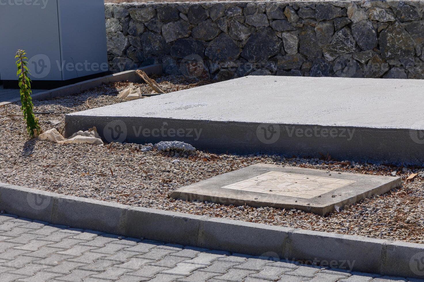 A newly paved section with gravel surrounding a square manhole cover near a stone wall on a sunny afternoon in a quiet neighborhood photo