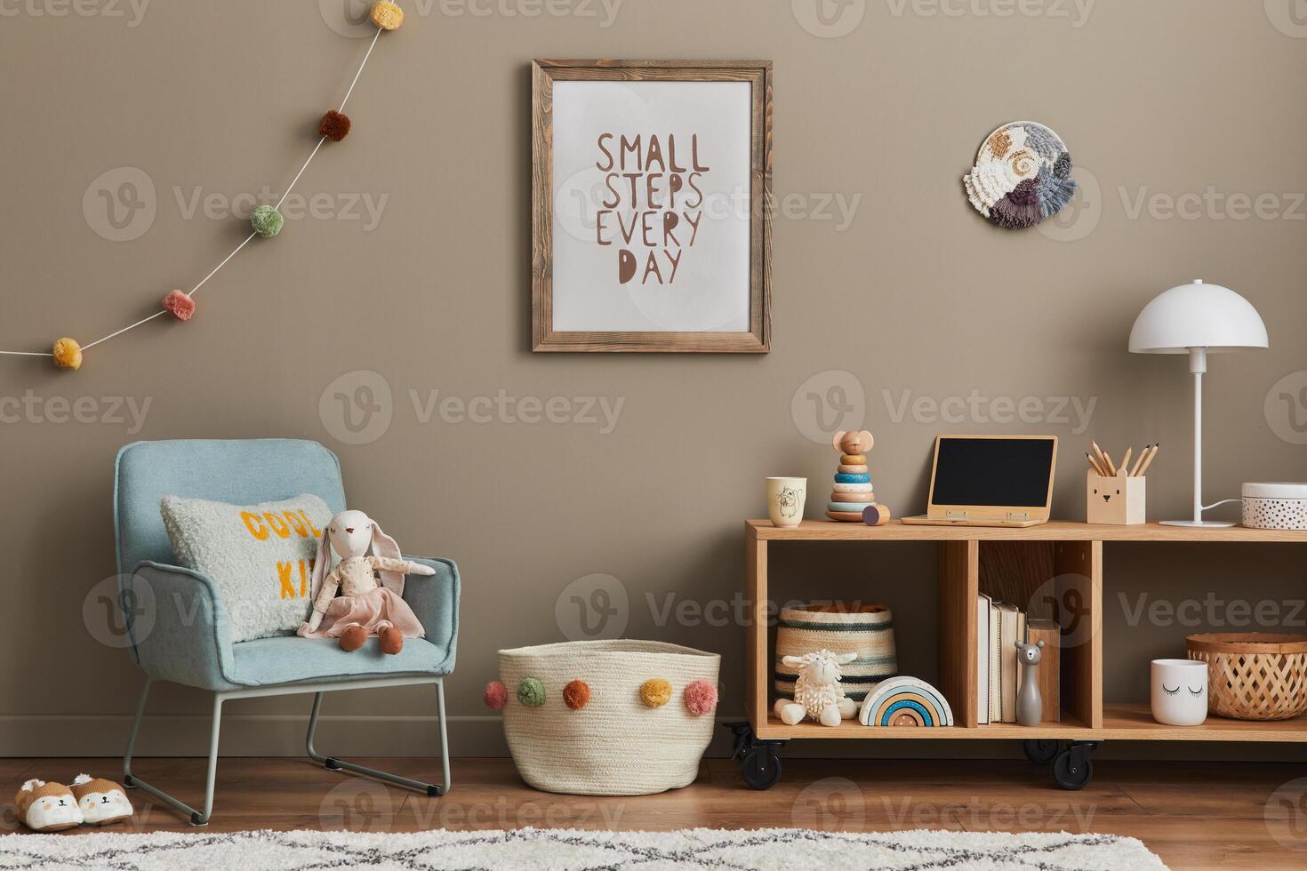 Cozy interior of child room with mint armchair, brown mock up poster frame, toys, teddy bear, plush animal, decoration and hanging cotton colorful balls. Beige wall. Warm kid space. photo