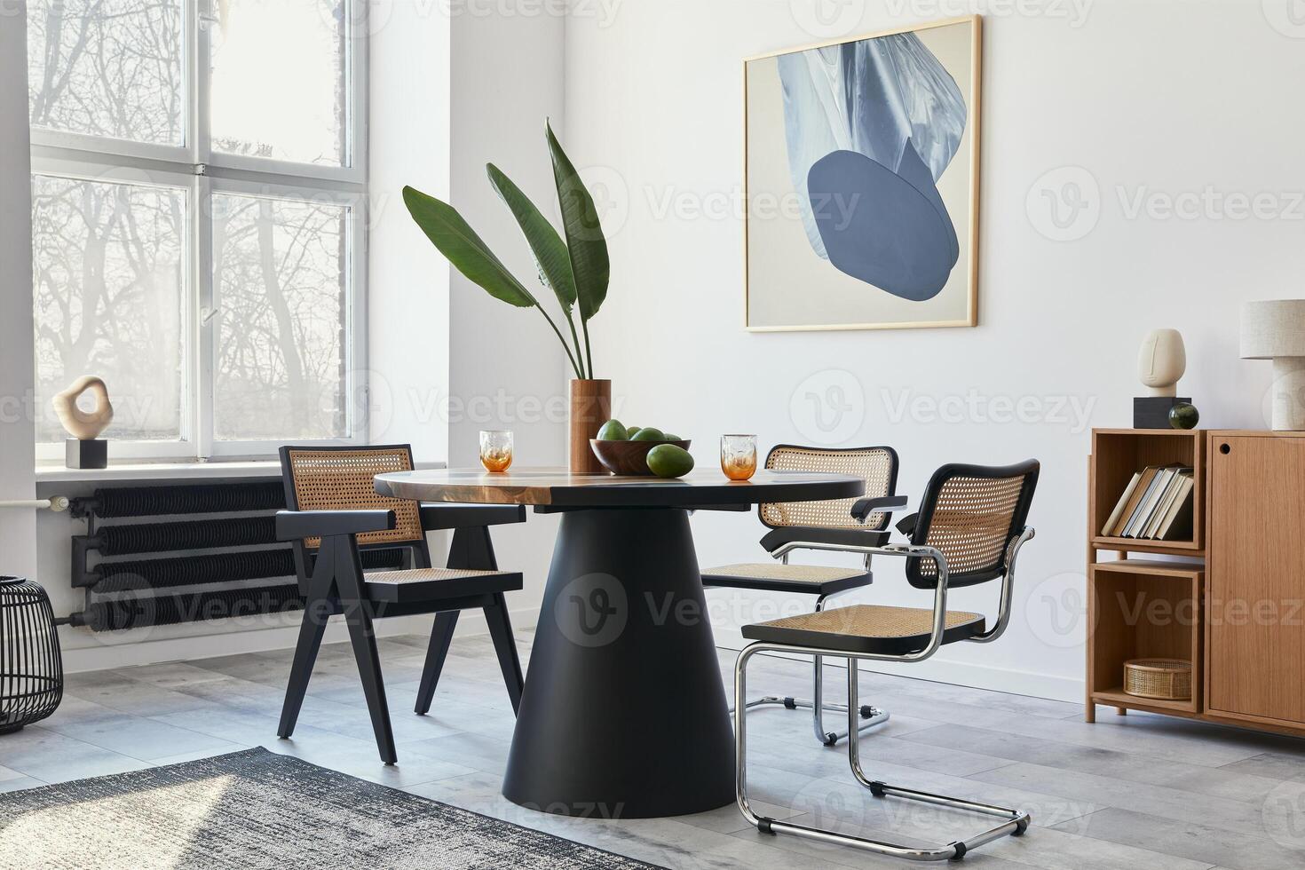 Stylish composition of dining room interior with design table, modern chairs, tropical leaf in vase, fruits, bookcase, abstract mock up paintings and elegant accessories in home decor. photo
