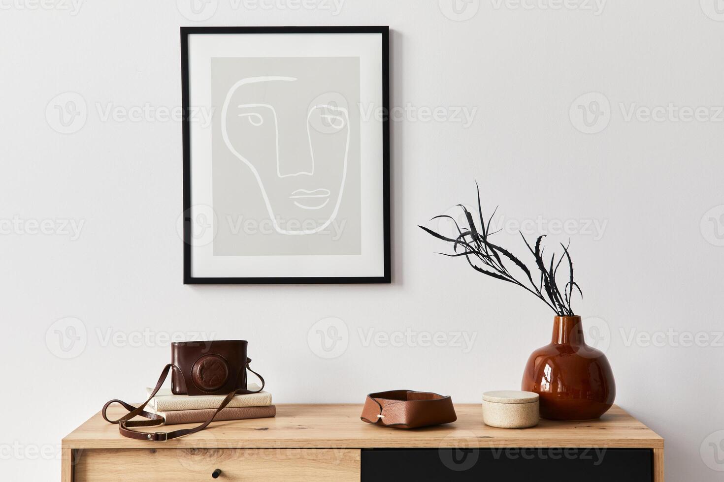 Stylish interior of living room with poster frame, wooden commode, book, leaf in ceramic vase, photo camera and elegant personal accessories. Minimalist concept of home decor.