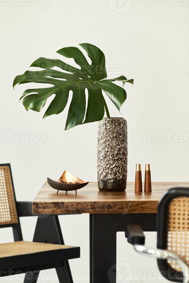 Modern concept of dining room interior with wooden table, chairs, plate with nuts, salt and pepper shaker and tropical leaf in vase. White wall. photo