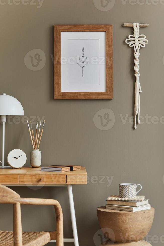 Stylish bohemian interior of home office space with wooden desk, rattan armchair, brown mock up poster frame, macrame, lamp, decoration and elegant personal accessories in home decor. photo
