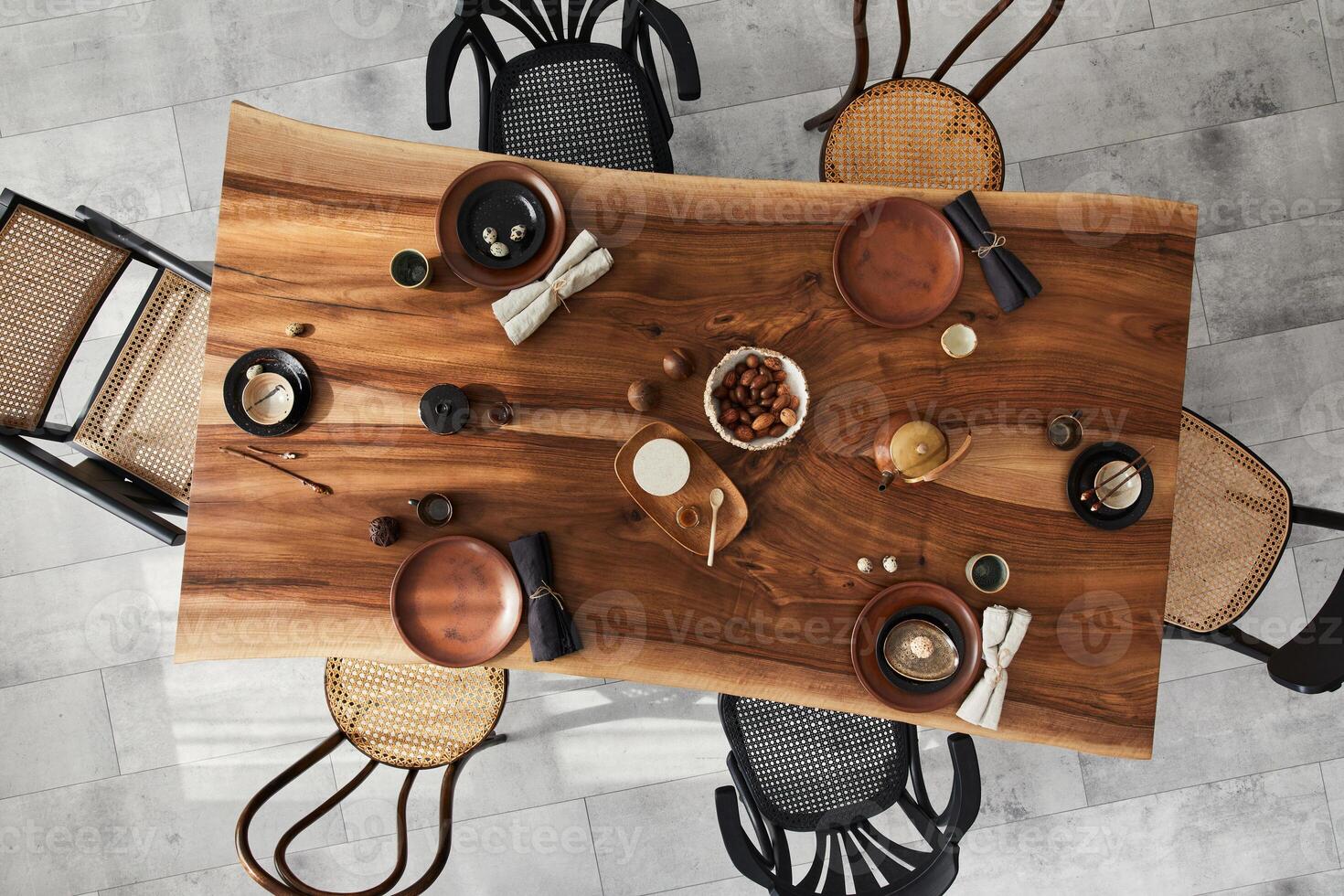 Stylish interior design of dining room with wooden walnut table, retro chairs, tableware, plates, teapot, food, decoration and elegant accessories. Cement floor. Template. Top view. photo