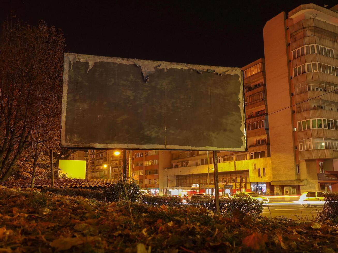 Empty billboard in urban area at night surrounded by illuminated buildings and fall leaves. photo