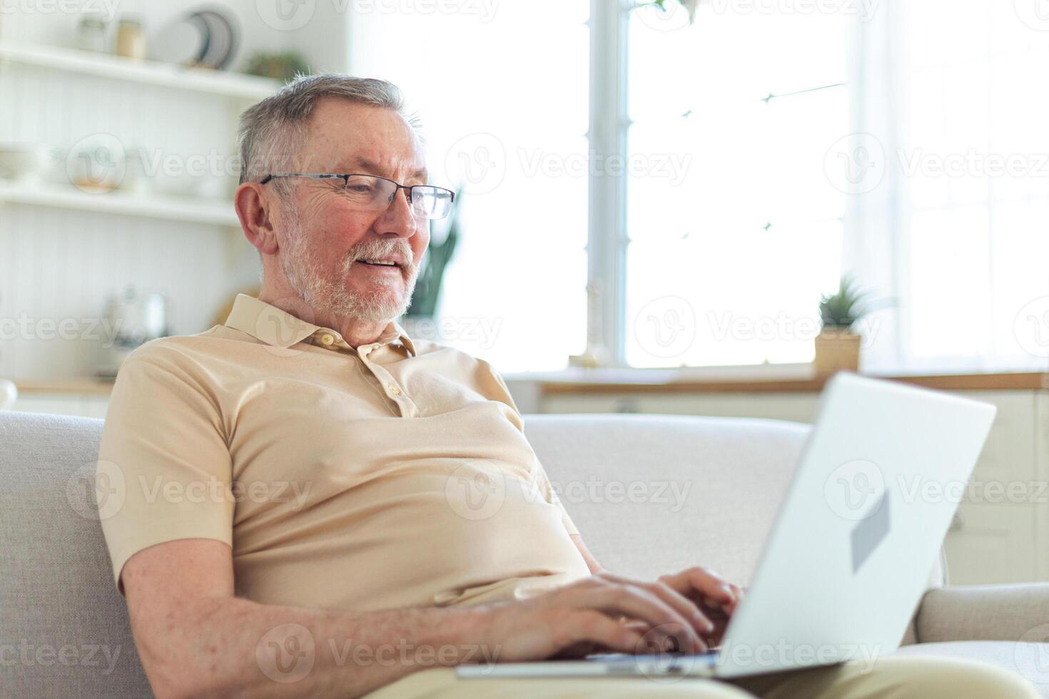 Confident stylish happy middle aged senior man using laptop at home. Stylish older mature 60s beard grandfather sitting at couch looking at computer screen typing chatting reading writing email photo