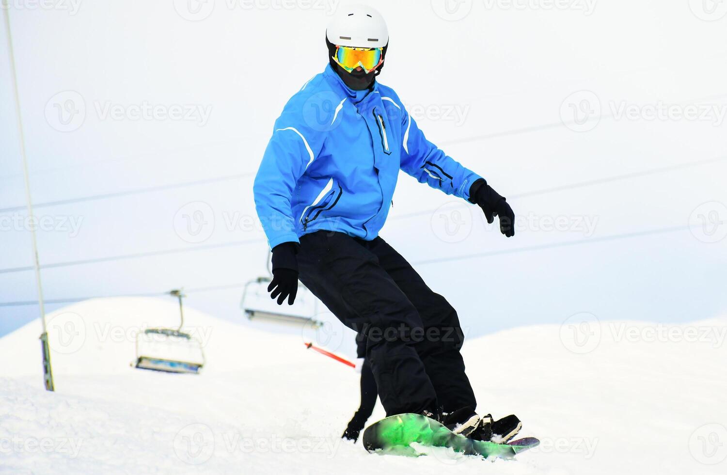 close up casual snowboard rider in blue black outfit looks down to camera isolated in white snowy background. Winter vacation with sports mountains photo