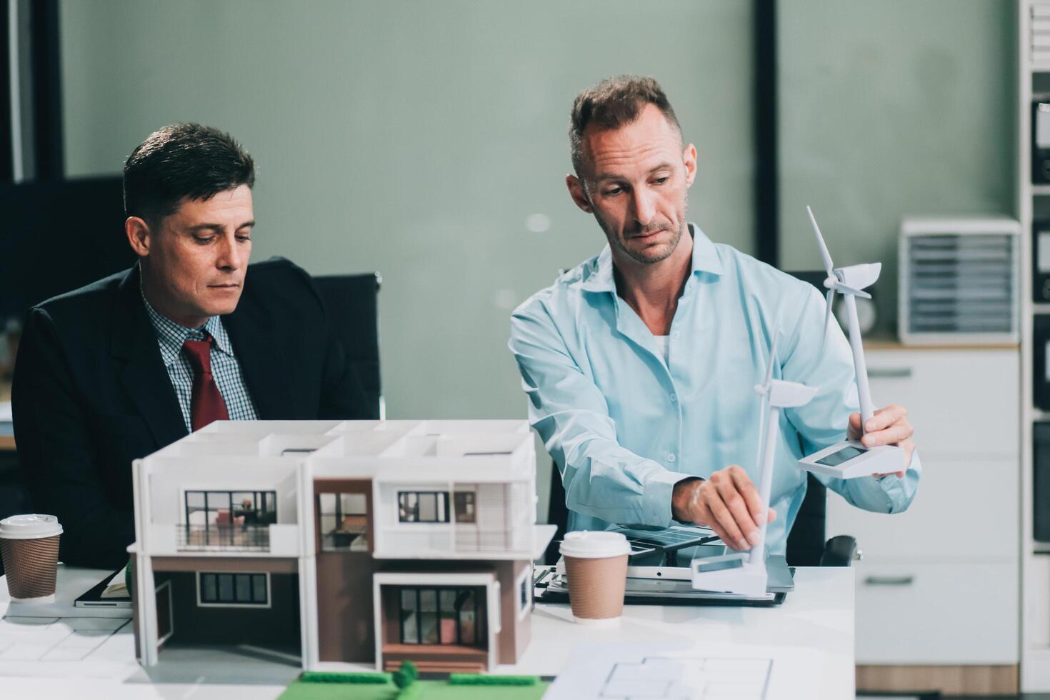 Engineers meeting about wind turbine model and house model. Renewable energy and housing development concept. Team of architects and engineers in modern office discussing design structure. photo