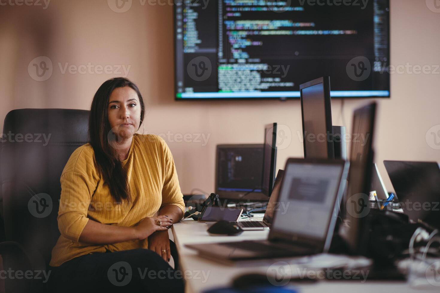 A businesswoman sitting in a programmer's office surrounded by computers, showing her expertise and dedication to technology. photo