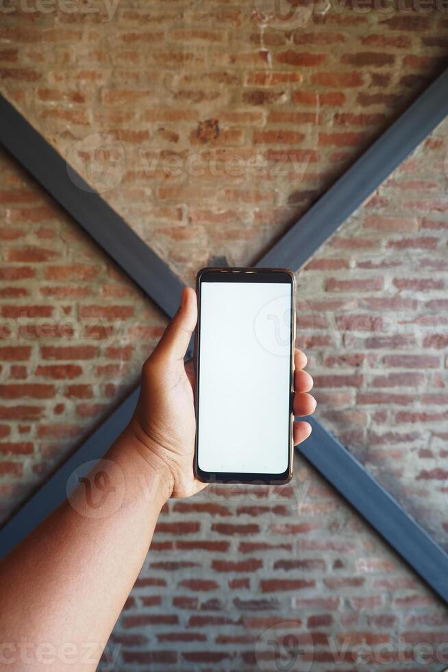 A hand holds a blank smartphone against a brick wall, offering a modern backdrop for branding photo