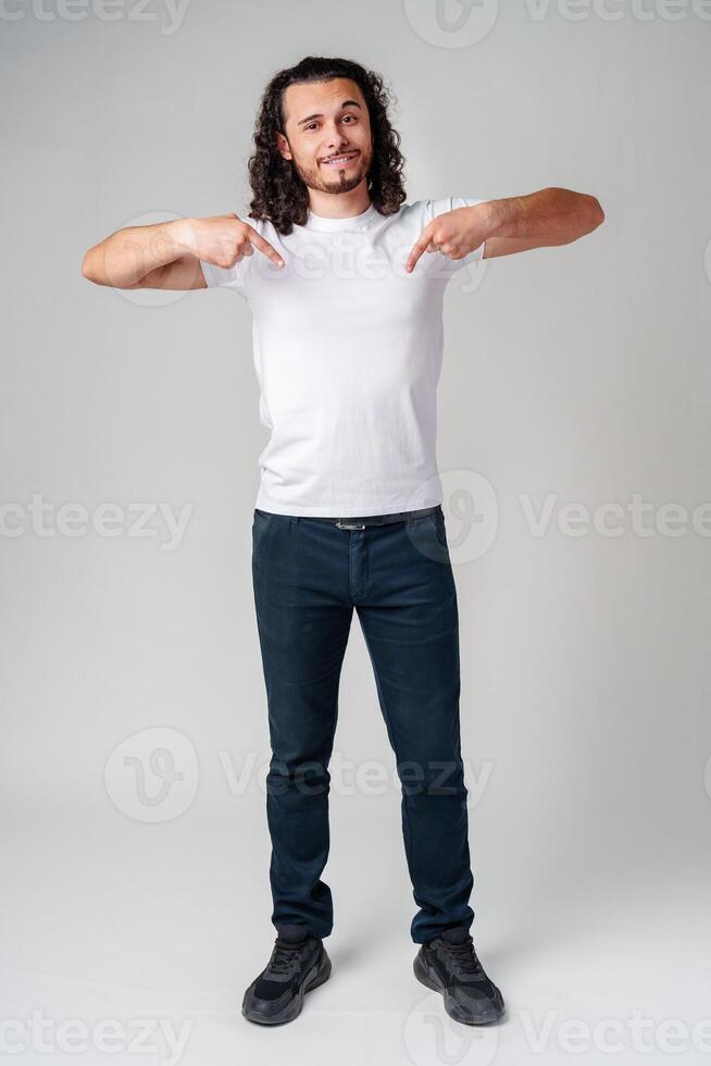 A young man with curly hair joyfully pointing down while wearing a plain white t-shirt against a neutral background photo