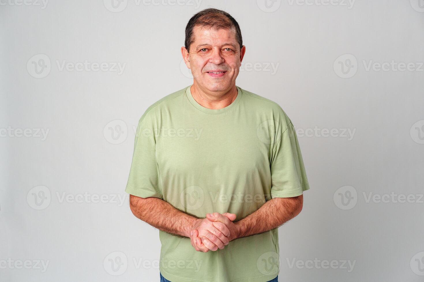 Middle-aged man smiling warmly while standing with hands clasped in front of light gray background photo