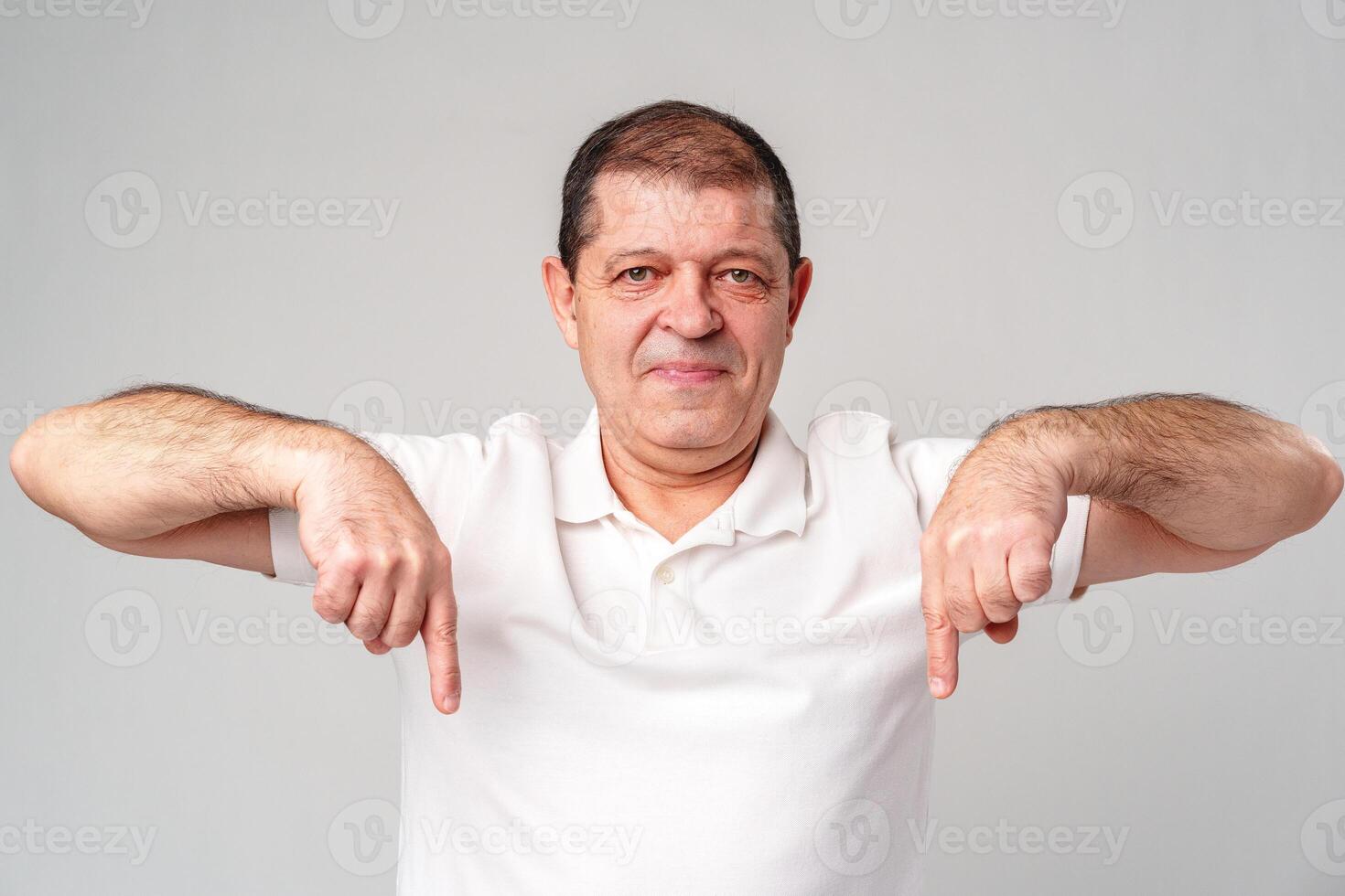 A man in a white shirt smiling and gesturing downwards in a studio with a light gray background photo
