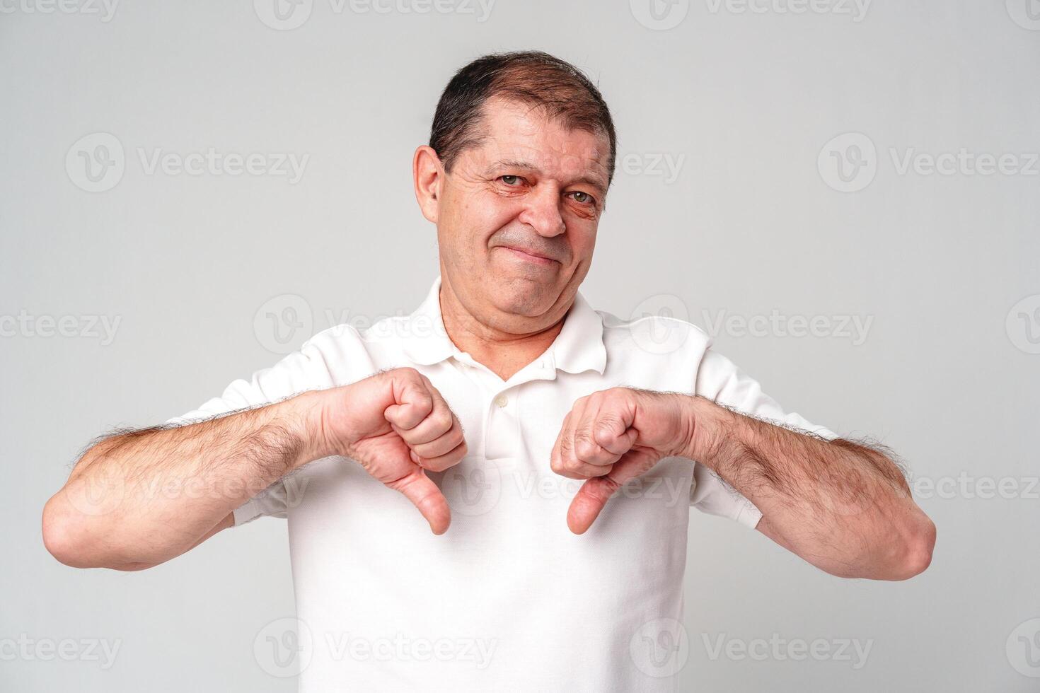 Older man expressing disapproval with thumbs down gesture while standing against a plain background photo