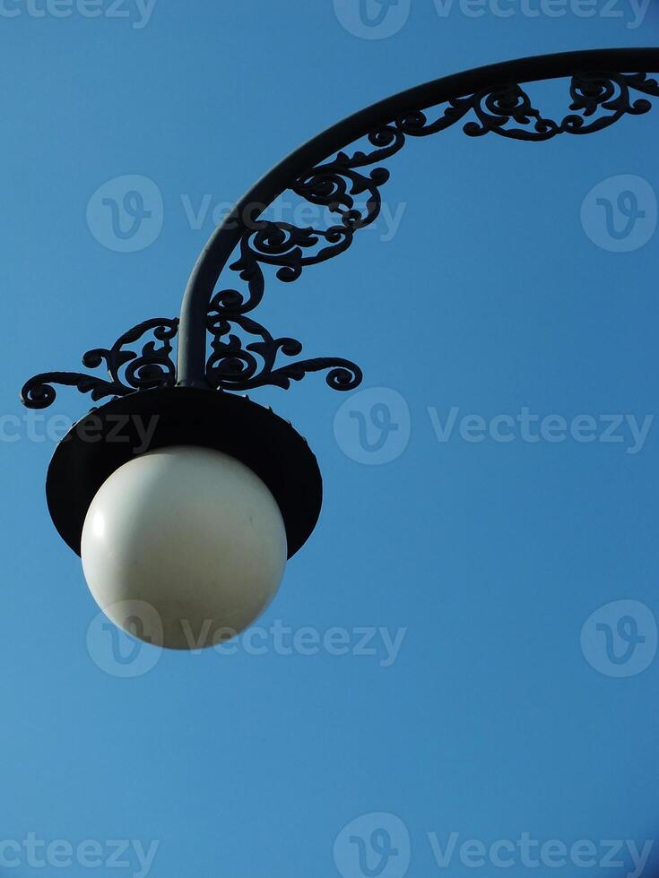 A street light with a white bulb is lit up against a blue sky. The light is on a metal pole with a decorative design. The scene is peaceful and serene photo