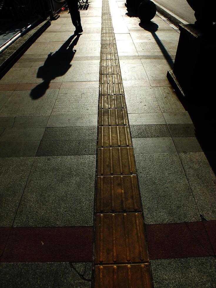 A person is walking on a sidewalk next to a bench. The bench is empty. The sidewalk is lined with bricks and has a shadow cast by the person photo