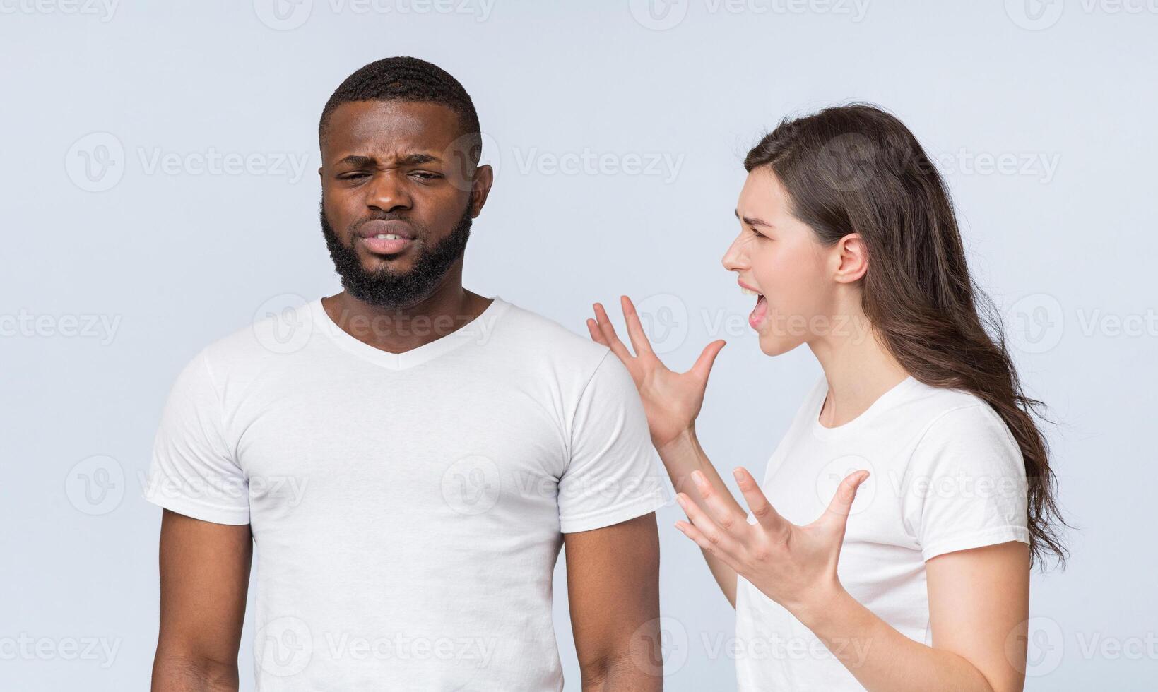 Furious young woman screaming at her annoyed black boyfriend, emotionally shouting and gesturing over light background photo