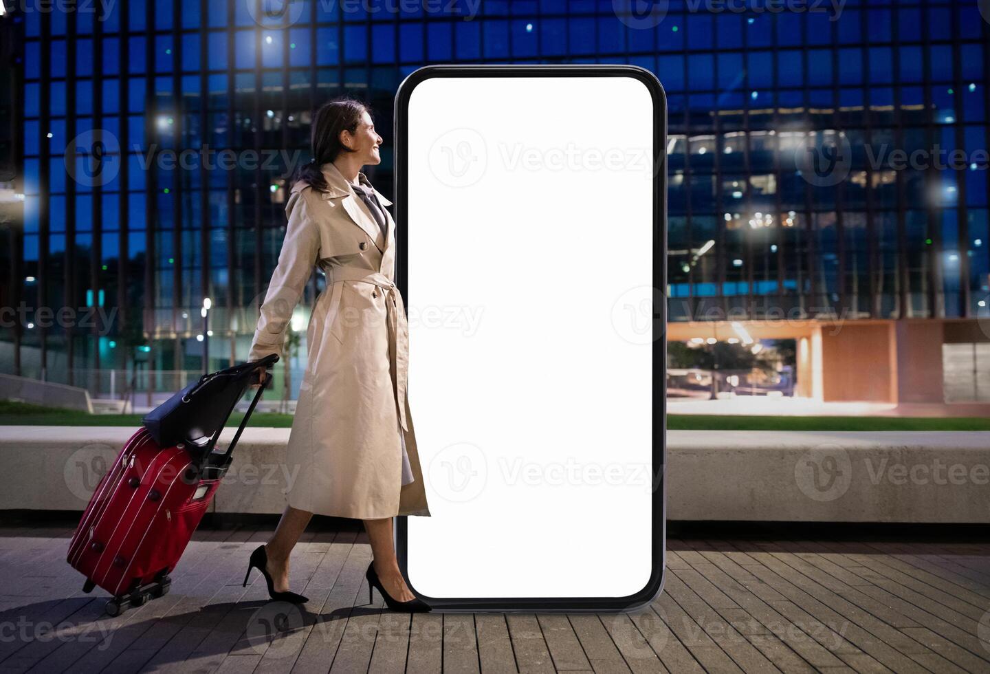 A woman carrying a red suitcase strolls by a massive blank smartphone screen in an urban setting during evening hours. The lights from nearby buildings create a vibrant atmosphere. photo