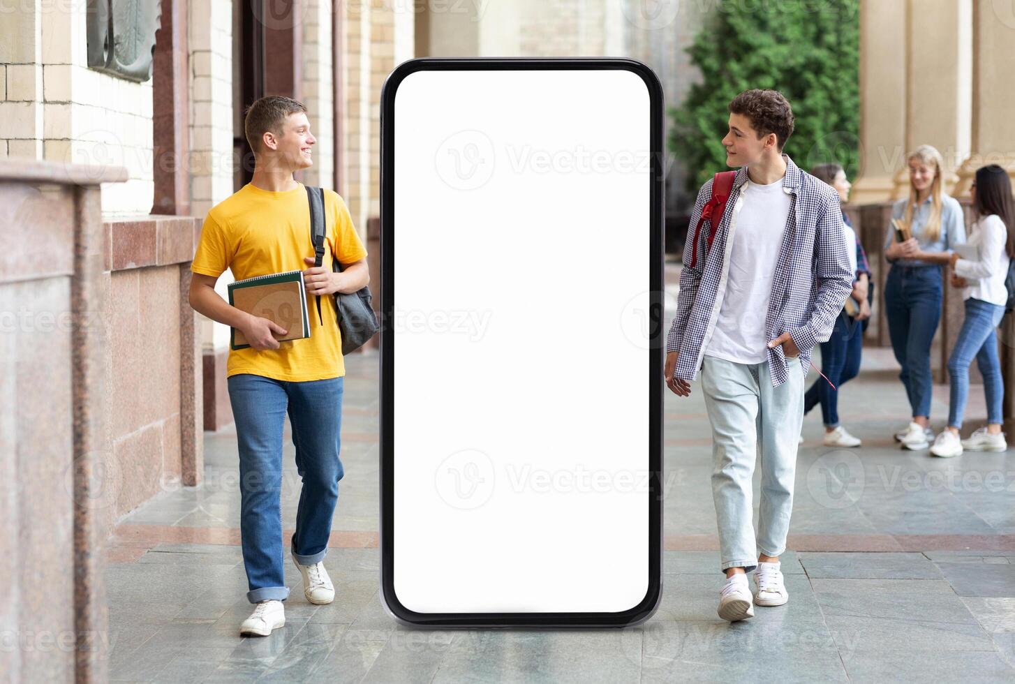 Two young men stroll through a school corridor, one holding books while the other carries a backpack. A large blank mobile screen is prominently displayed nearby. photo