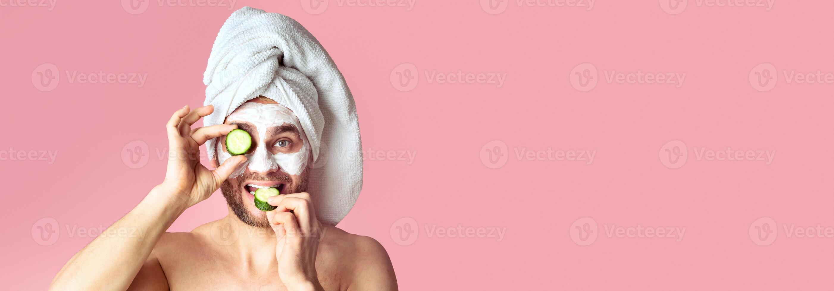 Millennial man makes face mask and eats cucumber on pink background photo