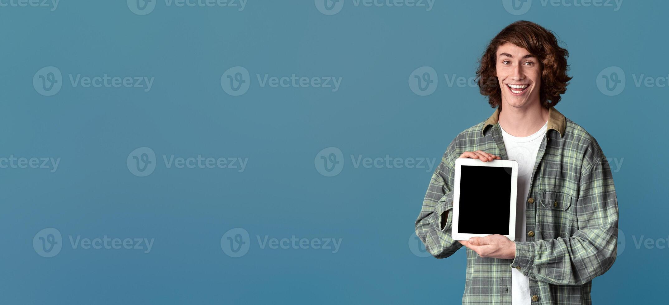 Curly millennial guy holding tablet computer with blank screen on color background, mockup for your design photo
