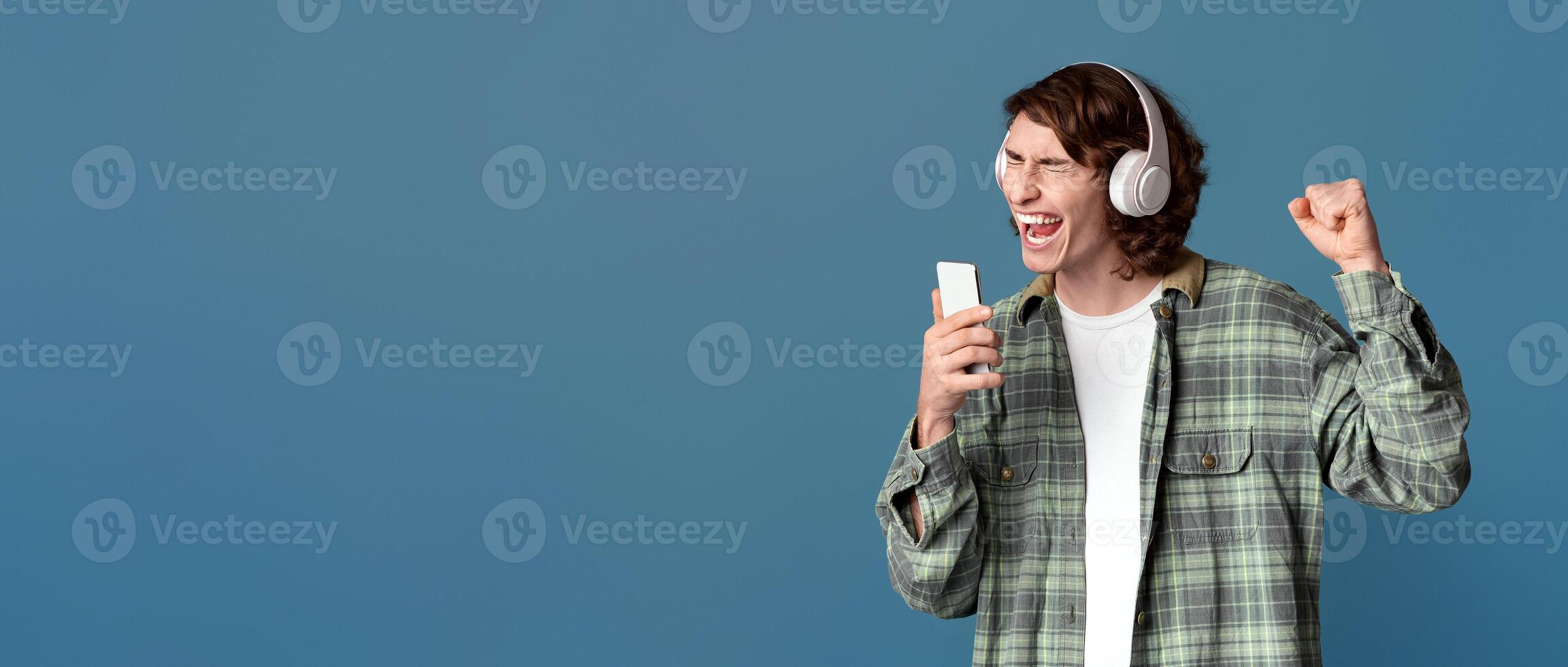 Trendy teenager in headset using smartphone as mike, singing along with his favorite song, color background. Copy space photo