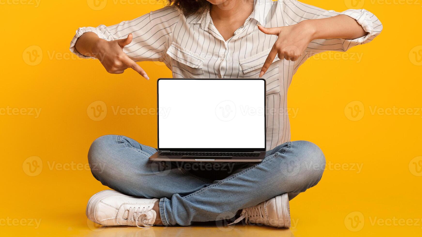 Unrecognizable black girl pointing at laptop with blank black screen at her laps, sitting on floor over yellow background in studio, panorama, crop photo