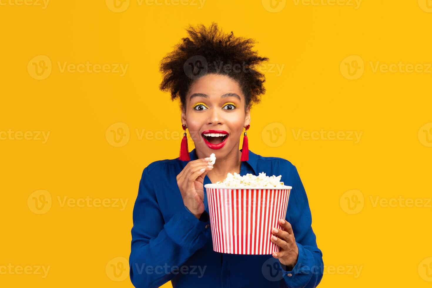 Free Time. Portrait of excited black girl eating popcorn and looking at camera, watching movie isolated over yellow wall photo